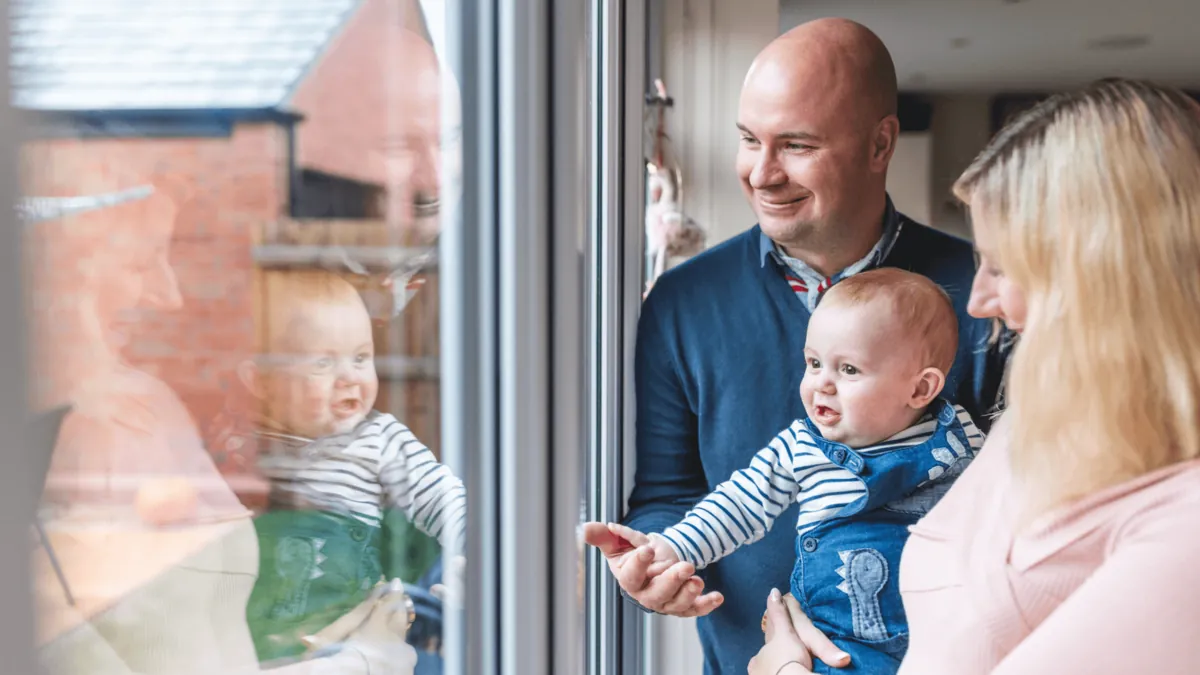 Parents holding a baby, looking out through a large new window from inside the house